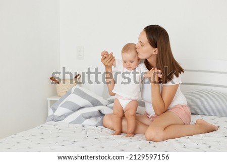 Image of lovely mother, attractive woman wearing white shirt and short sitting on bed with her toddler daughter, mommy kissing her child, expressing love and gentle.