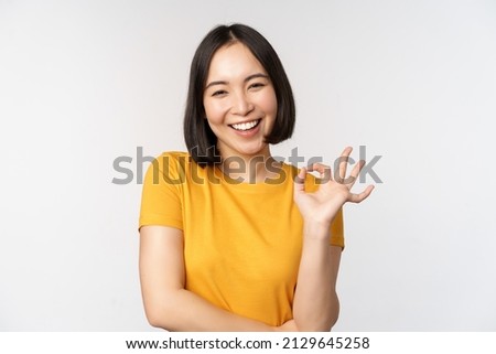 Beautiful young woman showing okay sign, smiling pleased, recommending smth, approve, like product, standing in yellow tshirt over white background