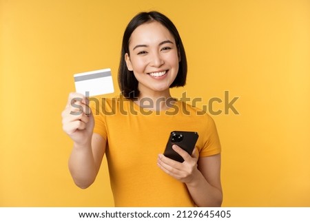 Joyful asian girl smiling, showing credit card and smartphone, recommending mobile phone banking, standing against yellow background