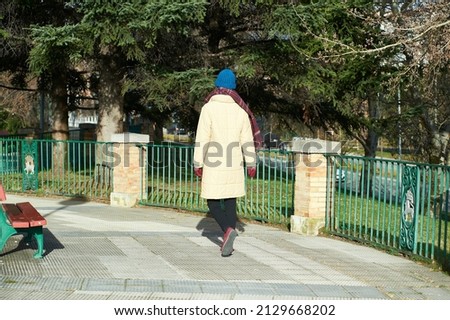 woman in an off-white coat with gloves and a blue wool hat walks along the sidewalk next to a red wooden bench