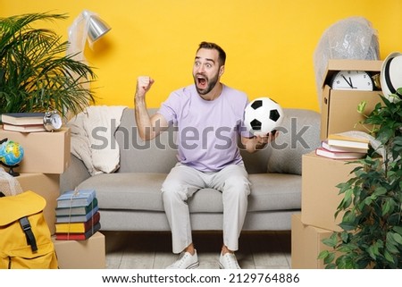 Crazy owner man expressive gesticulating support favorite team with ball sits in living room on sofa at home unpacking stuff rents flat isolated on yellow wall. Relocation moving in apartment concept