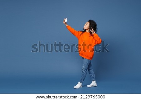 Full length of cheerful young african american woman in casual bright orange sweatshirt standing doing selfie shot on mobile phone showing victory sign isolated on blue background studio portrait