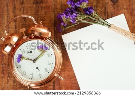 gold alarm clock with blue flowers instead of clock's hands and paper with small bouquet of flowers in the up on wooding surface. Spring time. Time for spring. place for text. topview
