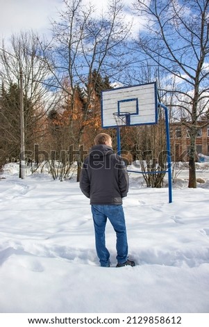 A man in a gray jacket and jeans stands near an old basketball hoop in winter. White snow, picturesque trees and blue sky with fluffy clouds. Vertical image.