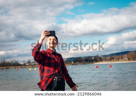Girl in a red plaid shirt with black short hair takes a selfie on a mobile phone on the river bank.