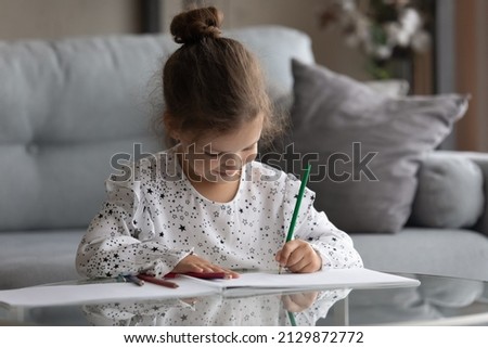 Happy adorable small preschool 6s child girl sitting at table in living room, enjoying drawing pictures with colored pencils in paper album, involved in creative hobby activity alone at home.