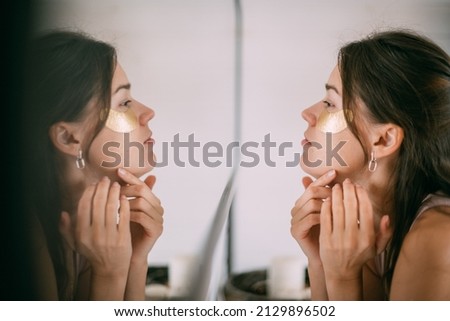 Young woman with patches under her eyes at the mirror at home. A beautiful brunette makes facial skin care procedures with hyaluronic patches near a large mirror in a bright room.