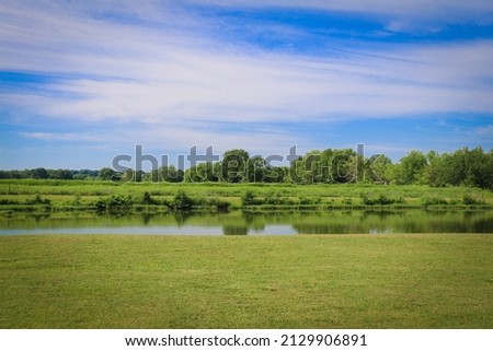 a rural lake green grass wild nature preserve park grassland pond reflection blue sky cloudscape Royalty-Free Stock Photo #2129906891