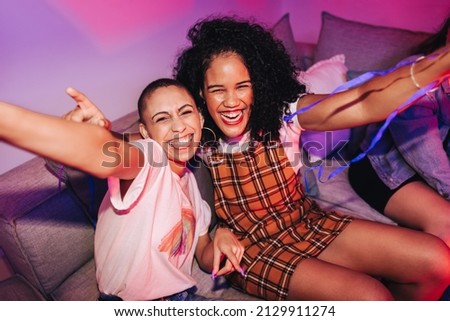 Taking a happy selfie at a house party. Two happy young women taking a picture together while sitting on a couch in neon light. Best friends having a good time together on the weekend.