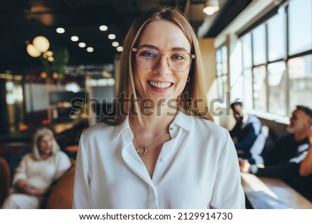 Successful young businesswoman smiling at the camera in a co-working space. Happy young female entrepreneur standing in a modern workplace with her co-workers in the background. Royalty-Free Stock Photo #2129914730