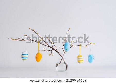 Minimalistic Easter still life. A glass vase with a branch on which colorful Easter eggs hang on a white background. Interior photography.