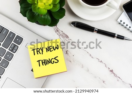 Try new things phrase on yellow adhesive note on top of laptop on office desk, pen and potted plant