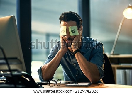 No time to sleep. Shot of a tired young businessman working late in an office with adhesive notes covering his eyes.