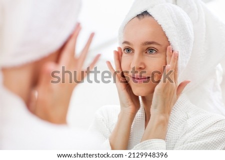 Happy 40s middle aged woman model touching face skin looking in mirror reflection. Smiling adult lady pampering, healthy moisturized skin care, aging beauty, skin care treatment cosmetics concept Royalty-Free Stock Photo #2129948396