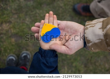 hand of a child in the hand of a man in camouflage with a yellow and blue heart. Family, patriotism, unity, support. Russia's invasion of Ukraine, a request for help to the world community Royalty-Free Stock Photo #2129973053