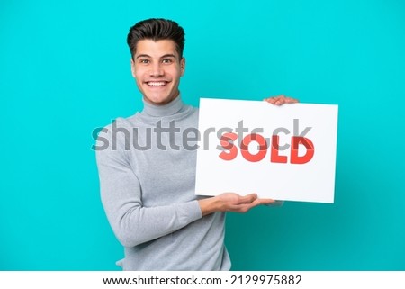 Young handsome caucasian man isolated on blue bakcground holding a placard with text SOLD with happy expression