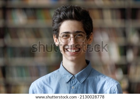 Head shot portrait of happy handsome Jewish male student in eyeglasses posing at blurred library background. Smiling Z generation smart guy looking at camera, high school or college education concept.