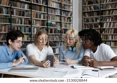 Focused smart four multiracial students using cellphone, web surfing useful information or watching educational video online, preparing together for exam in library, working on high school assignment. Royalty-Free Stock Photo #2130028844