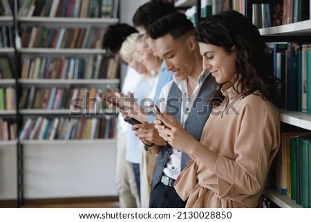 Side view addicted to modern tech happy young diverse students using cellphone apps, enjoying web surfing or communicating on social network standing near bookshelves in college library, copy space.