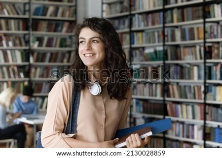 Happy dreamy confident Hispanic female college student with headphones on neck and educational books in hands looking away, thinking of future career or university graduation exams in library.
