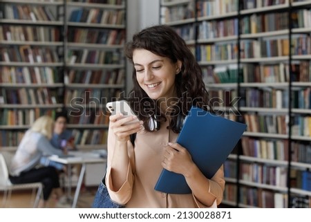 Joyful Z generation beautiful curly Hispanic 20s girl with folder in hands looking at smartphone screen, reading message with good news, communicating in social network or web surfing in library.