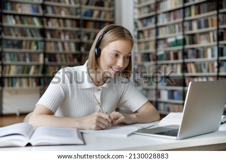 Happy young beautiful female student wearing headphones, enjoying watching educational lecture on computer, reading books doing homework or college project, studying sitting at table alone in library Royalty-Free Stock Photo #2130028883