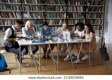 Full length concentrated team of multiethnic millennial college university students studying sitting together at big table, working on project in library using gadgets and paper books, writing notes. Royalty-Free Stock Photo #2130028886