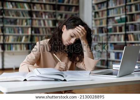 Unhappy stressed young Hispanic female student feeling tired preparing for examination, reading books, web surfing information, writing notes in copybook, overworking in modern college library. Royalty-Free Stock Photo #2130028895