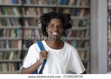 Head shot portrait of smiling 20s African American guy with backpack posing near bookshelves in modern library. Profile photo of happy smart millennial multiethnic male student looking at camera.