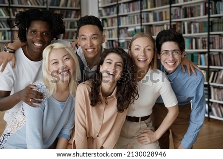 Portrait of bonding happy friendly young multiethnic college students embracing, posing in library near bookshelves. Group of joyful diverse millennial people looking at camera, relations concept.