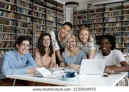 Portrait of joyful six multiethnic millennial smiling friends enjoying preparing for exams or common college project, sitting together at table in modern library, using computer and textbooks. Royalty-Free Stock Photo #2130028955