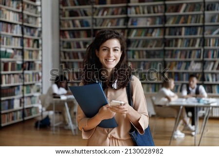 Portrait of smiling Hispanic female college student with headphones in neck holding folder and cellphone in hands, using different modern tech gadgets in public library, browsing internet, web surfing