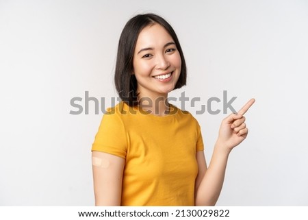 Covid-19 and vaccination concept. Smiling asian girl with band aid on shoudler, pointing finger at banner, showing vaccine campaign, standing over white background