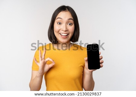 Excited asian girl showing mobile phone screen, okay sign, recommending smartphone app, standing in yellow tshirt over white background
