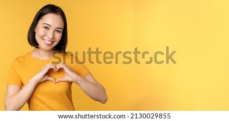 Beautiful asian girl showing heart, love gesture and smiling white teeth, express care and sympathy, standing over yellow background