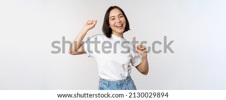 Happy dancing korean girl posing against white background, wearing tshirt Royalty-Free Stock Photo #2130029894