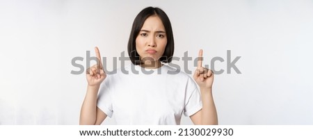 Sad and disappointed young asian woman pointing fingers up, showing advertisement with upset face expression, white studio background