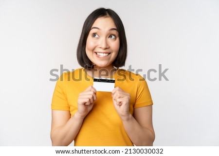 Portrait of beautiful korean girl holding credit card, recommending bank service, standing in yellow tshirt over white background