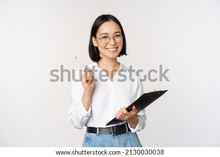 Smiling young asian woman taking notes with pen on clipboard, looking happy, standing against white background Royalty-Free Stock Photo #2130030038