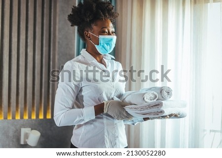 Maid with face protective mask cleaning a hotel room during pandemic. Maid working at a hotel wearing a protective face mask and bringing fresh clean towels in the room, during COVID-19