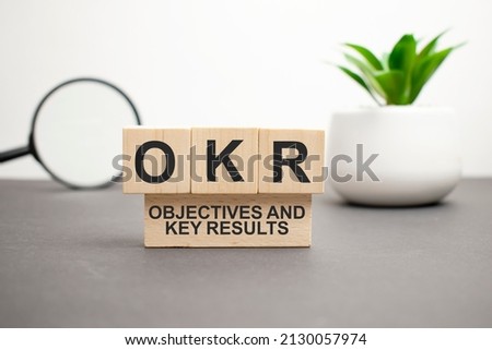 OKR word is written on wooden cubes on a gray background. close-up of wooden elements. In the background is a green flower. OKR - short for Objectives and Key Results