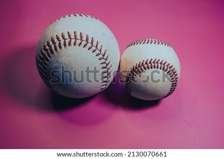 A softball and baseball next to each other, with pink background.