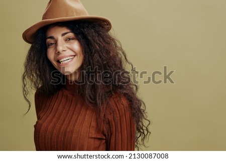 Close up portrait. Overjoyed stylish curly Latin female in brown hat, smiling at camera, laughing, saying Yeah, isolated green background. Copy space clothing fashion brands, free place for your ad. 