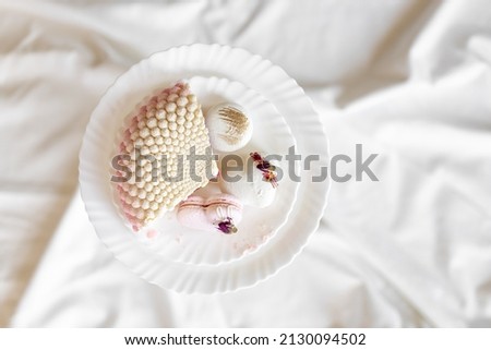 A white-pink piece of cake lies on a white plate on a light background. A pink heart-shaped cake lies on a white plate on a light background. Light macaroons on a white canvas. Delicious pastries