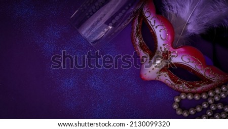 A masquerade mask, feathers, pearl beads and an empty champagne glass and sequins lie to the right on a dark purple background with copy space to the left, flat lay . Mardi gras celebration concept.