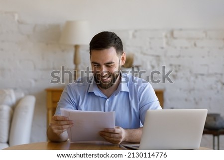 Handsome business man sit at table with laptop holding papers, read received contract, agreement, legal documents smile feel satisfied, got good news. Loan repayment, career advance, success concept Royalty-Free Stock Photo #2130114776
