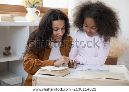 Caring young African American mother or nanny helping primary pupil cute little child daughter with school tasks, preparing homework together, learning writing on counting, sitting together at table. Royalty-Free Stock Photo #2130114821