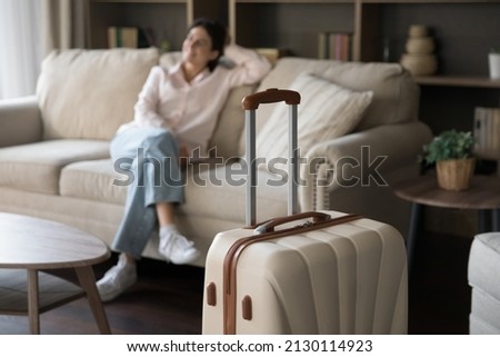 Focus on beige suitcase with blurred carefree young woman sitting on sofa on background. Joyful refreshed millennial Hispanic lady satisfied with vacation travel, relaxing alone in modern hotel room. Royalty-Free Stock Photo #2130114923