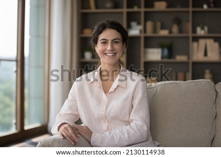 Portrait of happy confident millennial Hispanic female homeowner sitting on comfortable sofa at home, resting alone in modern living room. Web camera view smiling pretty young woman posing indoors.