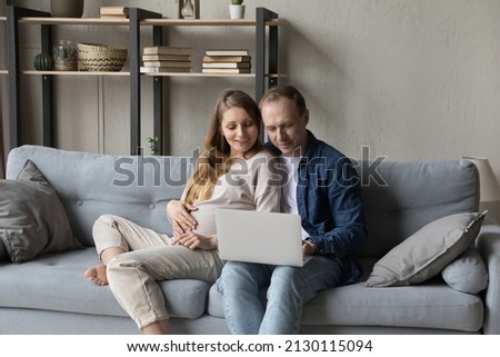 Happy family pregnant couple relaxing on comfortable couch together, using computer, shopping with online ecommerce app on laptop, browsing Internet, talking on video call, touching big belly Royalty-Free Stock Photo #2130115094